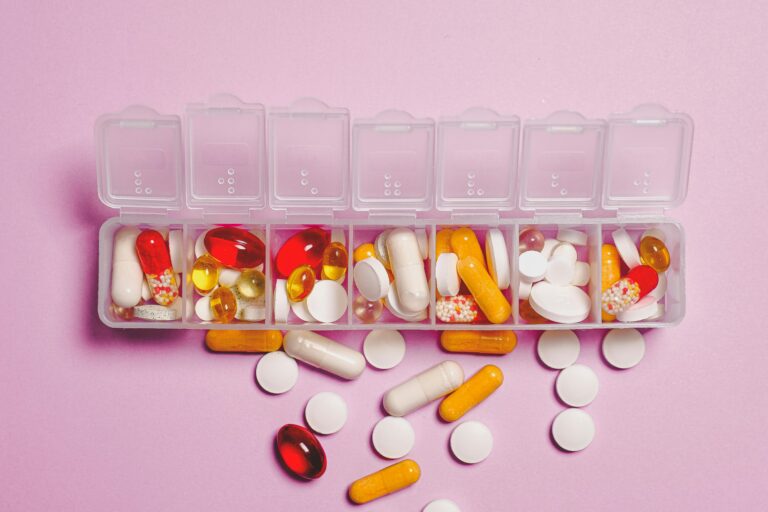 A variety of pills in a weekly pill organizer on a pink background.